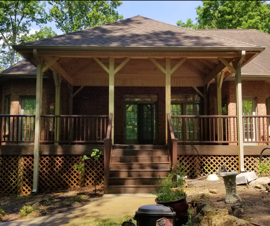 A brick house with a covered porch, wooden railings, and a brown roof. Steps lead up to a doorway.