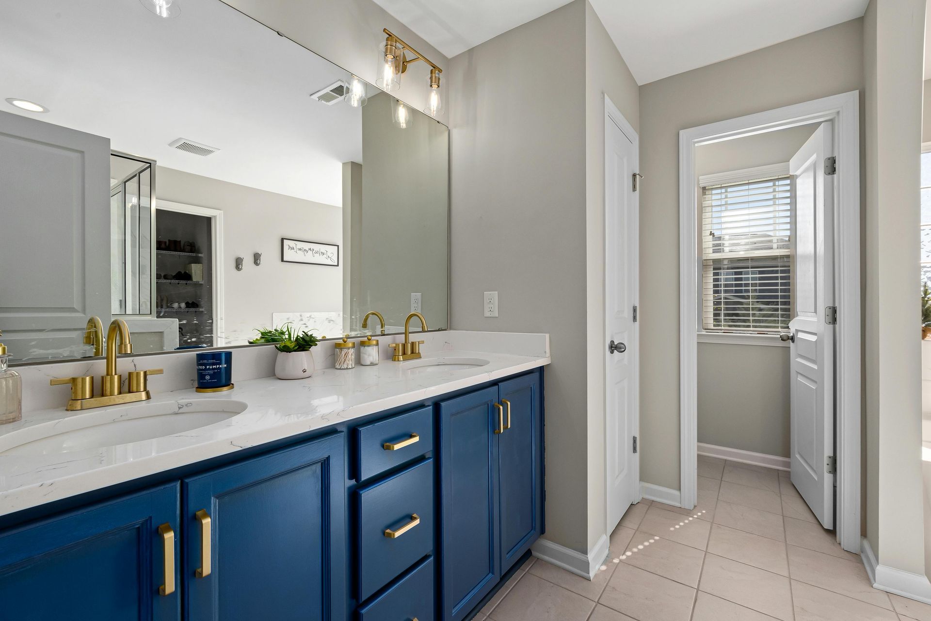 Blue vanity with gold fixtures and large mirror in a well-lit bathroom. A doorway leads to a bright hallway with a window.