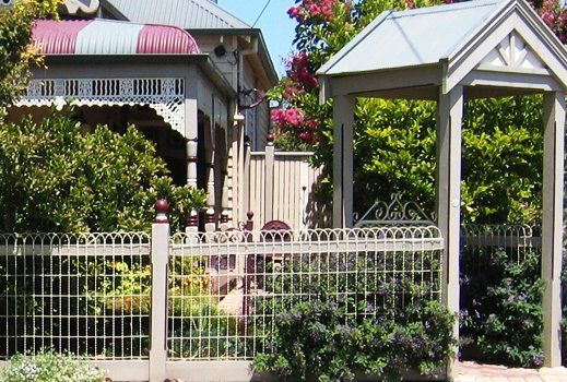 Ornate beige fence and gate in front of a Victorian-style house with a burgundy awning and a garden arbor.