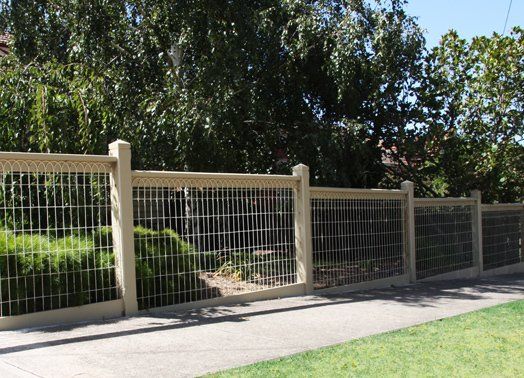 Beige fence with wire mesh panels bordering a sidewalk and lawn, with trees in the background.