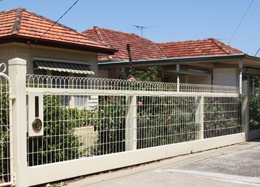 Cream-colored fence with gate in front of a house with orange tiled roof. Bushes behind the fence.