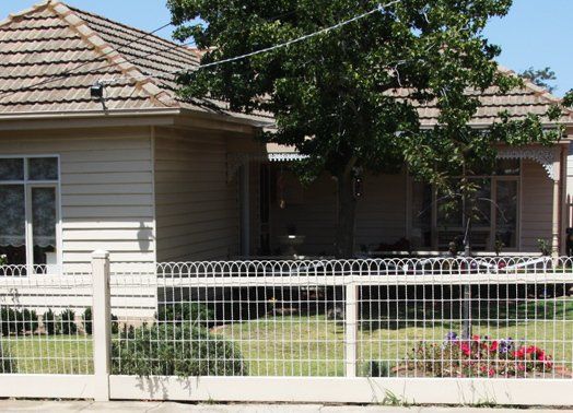 Tan house with tiled roof, surrounded by a white picket fence, a tree in front.