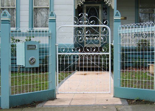White wrought-iron gate and fence, teal posts, leading to a light-colored walkway and house entrance.