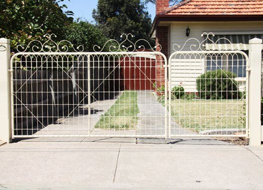Cream-colored ornate metal gates and fence in front of a house, driveway visible, with some greenery.