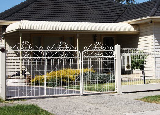 Cream-colored wrought iron gates in front of a beige house with a curved awning, green lawn, and dark roof.