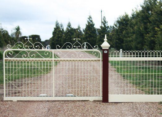 Cream-colored metal driveway gates, with ornate scrollwork, open onto a gravel driveway. A dark brown post is on the right.