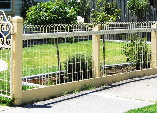 Cream-colored wire fence with matching posts, bordering a green lawn and sidewalk. Trees in the background.