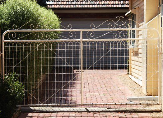 Cream-colored metal gate with decorative scrollwork, brick driveway, and a house facade in the background.