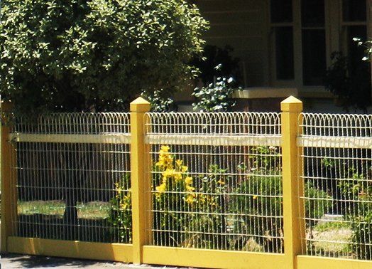 Yellow picket fence with wire mesh panels, fronting a garden with flowering plants.