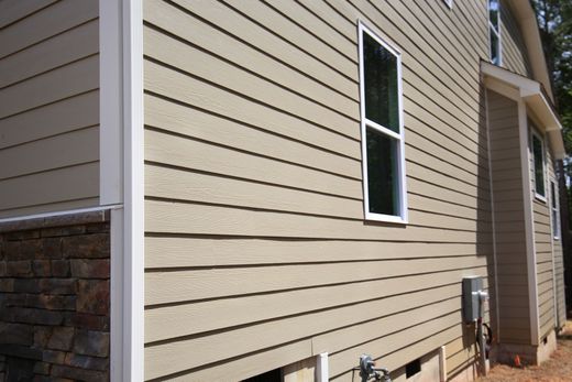 Beige vinyl siding exterior wall with a narrow window and utility meters beside a stone corner.
