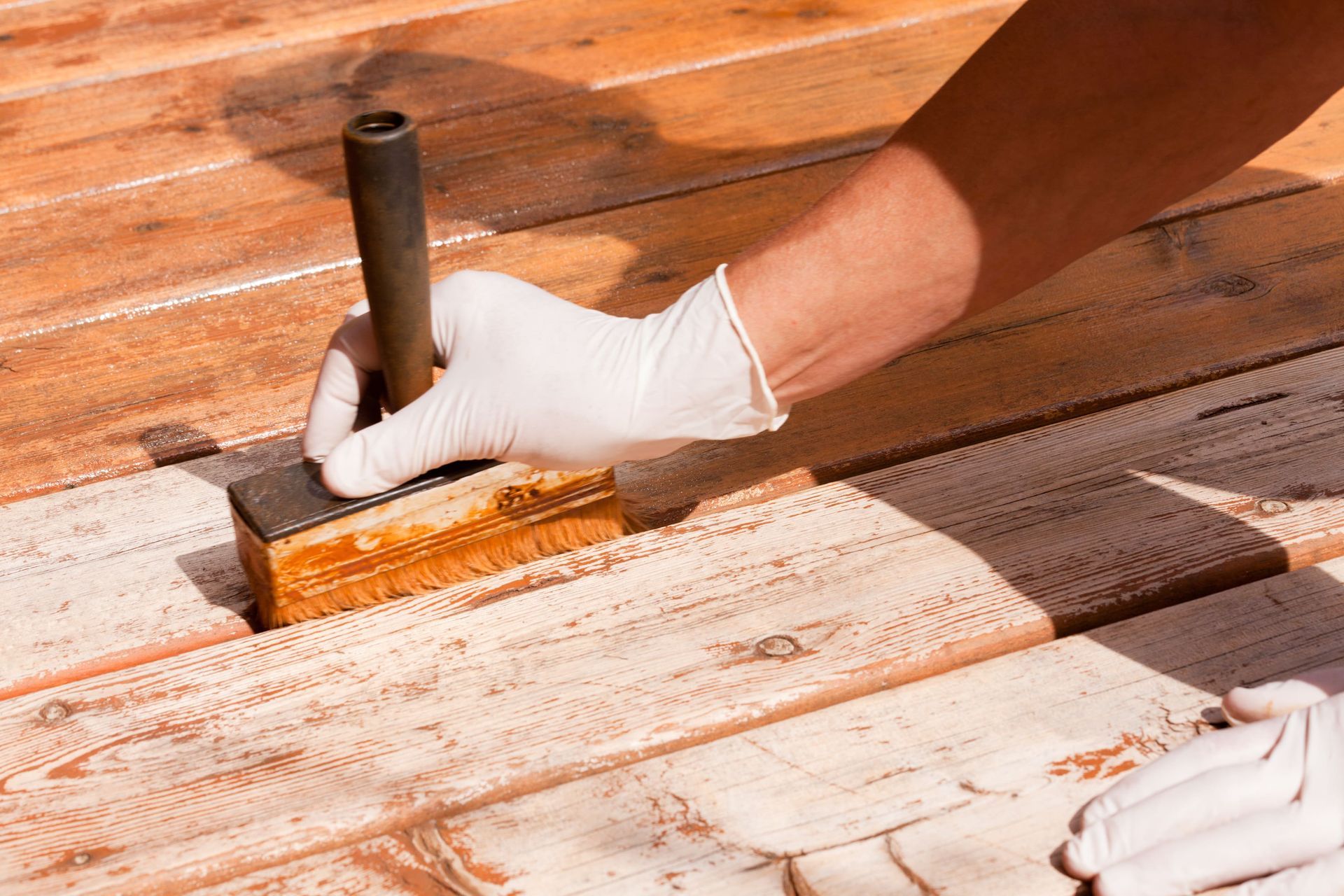 Gloved hand painting a white wooden deck with a brush and orange stain underneath