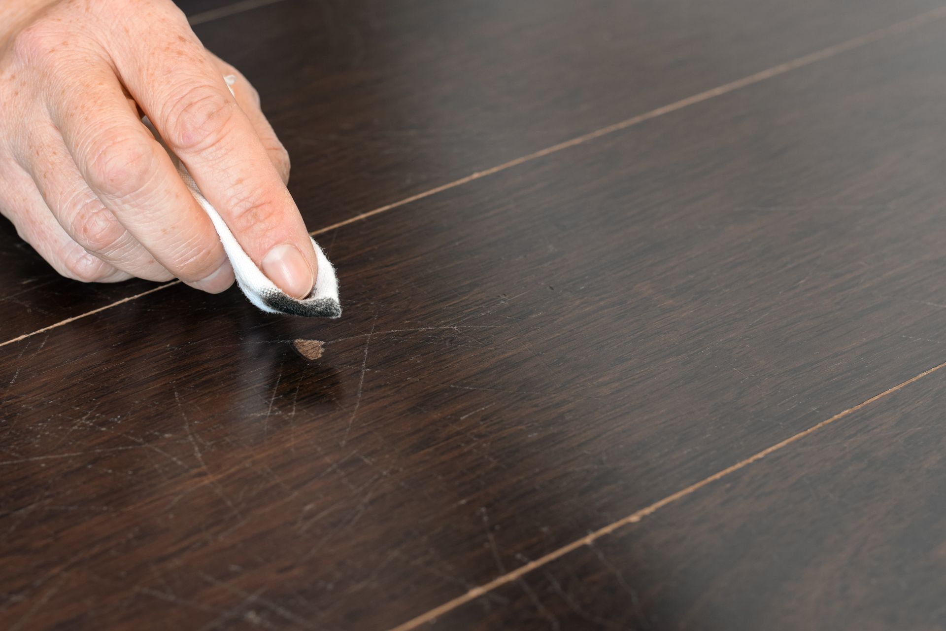 Hand rubbing chalk into a scratch on a dark wooden floor, concealing the mark