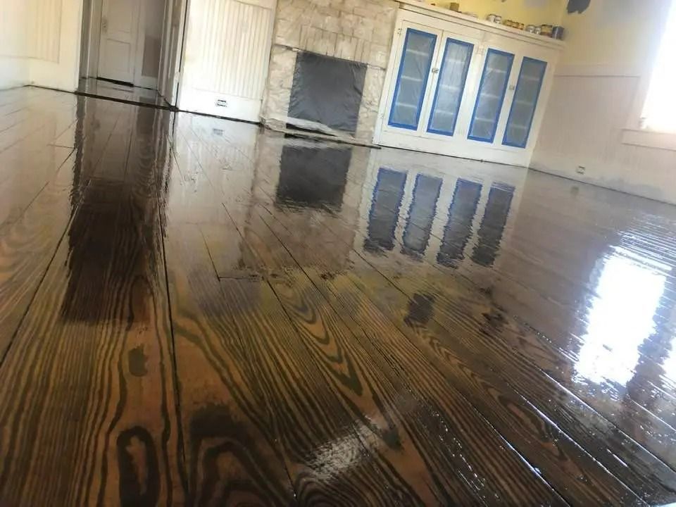 Flooded room with glossy wooden floor reflecting light, near a fireplace and windows; chairs line the wall.