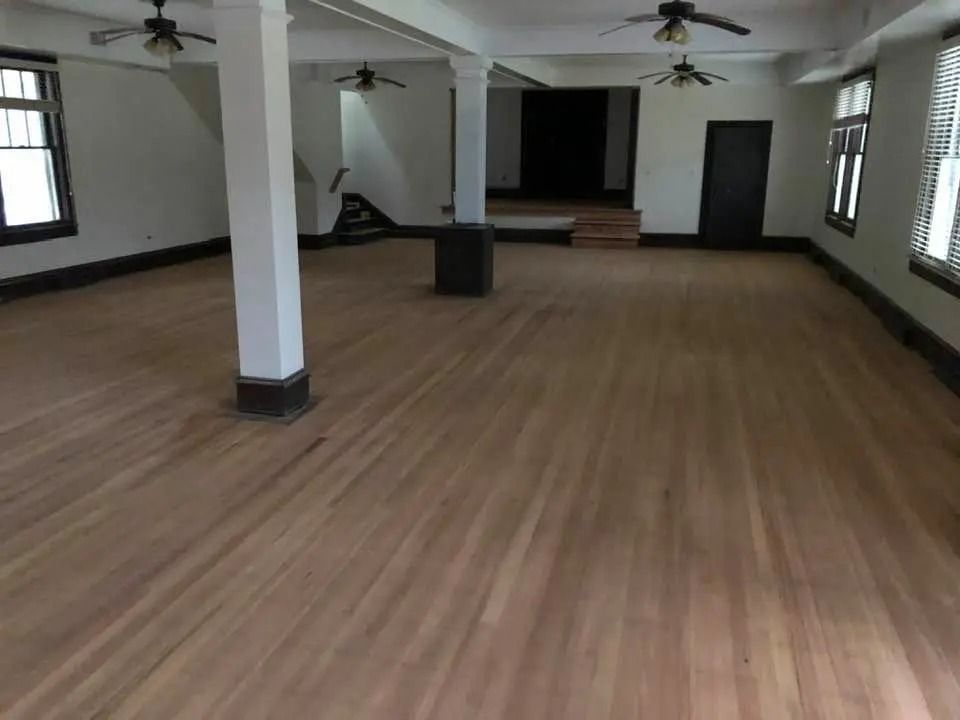 Empty hall with wood floors, white columns, ceiling fans, and windows along the walls.