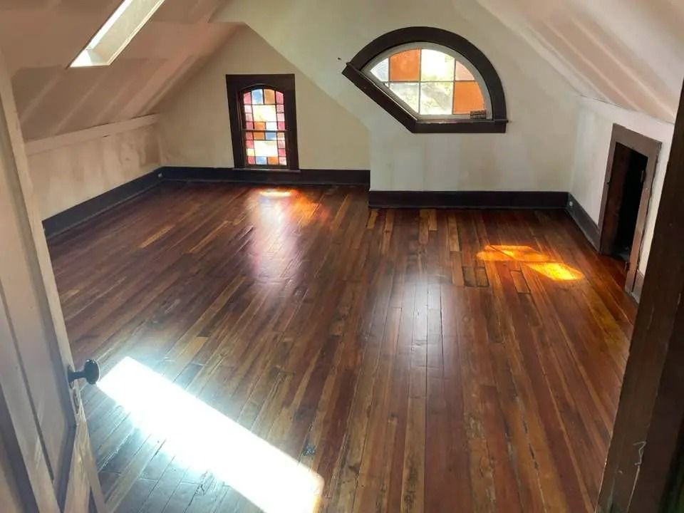 Empty attic room with dark wood floors, slanted ceiling, and small stained-glass windows.