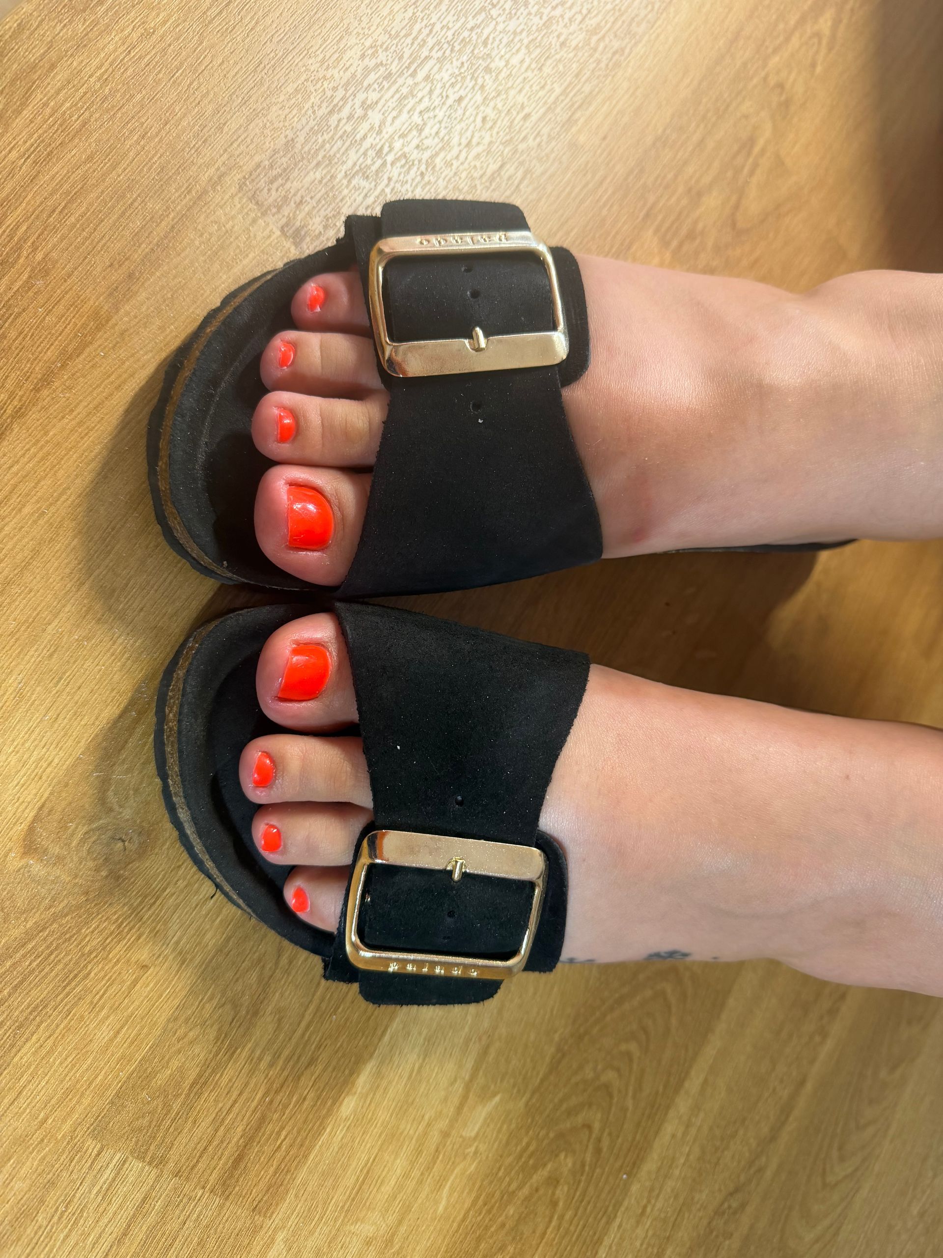 Füße in schwarzen Sandalen mit goldenen Schnallen und knallrotem Nagellack auf den Zehennägeln, auf einem Holzboden ruhend.
