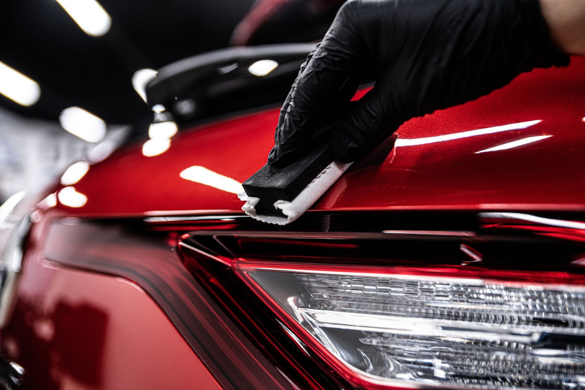 A hand in a black glove applies a ceramic coating with a small applicator block to a shiny red car body.