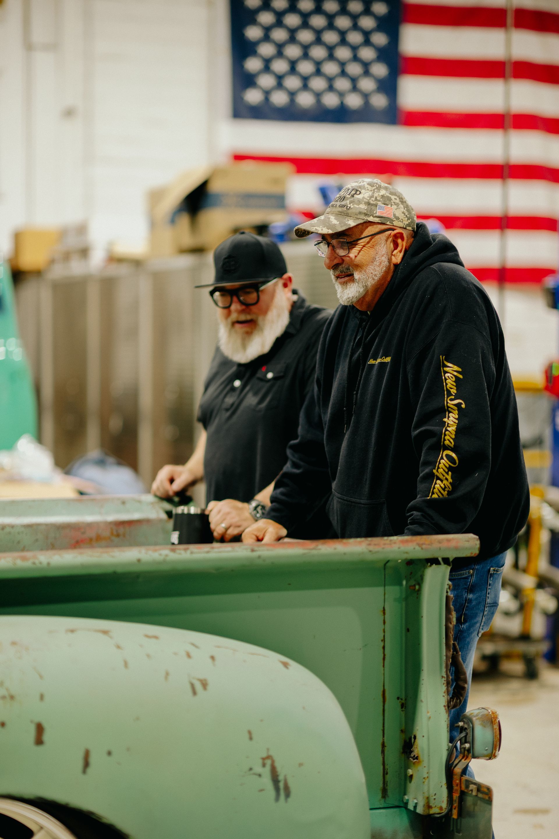 Two men in black jackets standing by machinery in a workshop with an American flag in the background