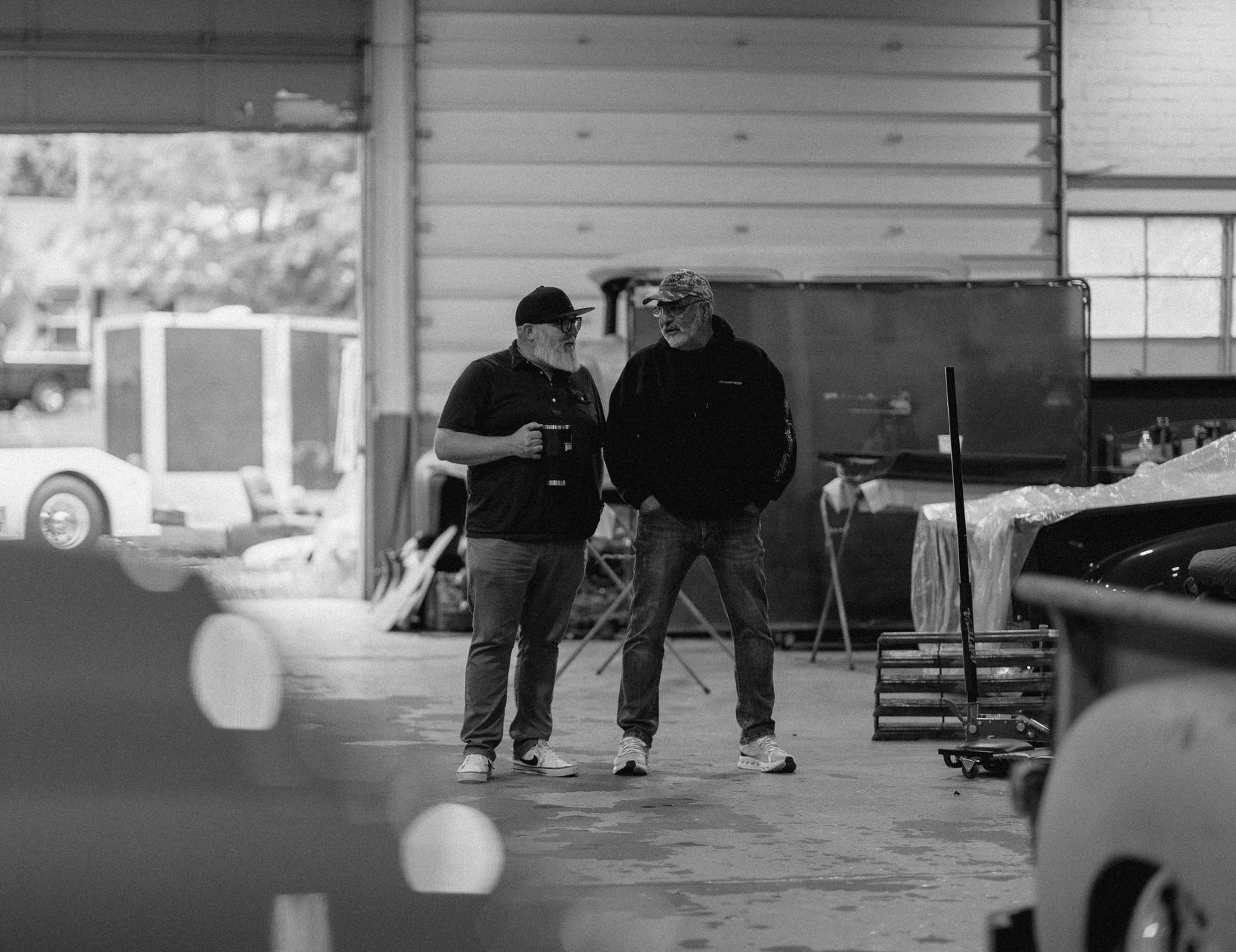 Two people walking in a garage, one gesturing while talking beside parked vehicles.
