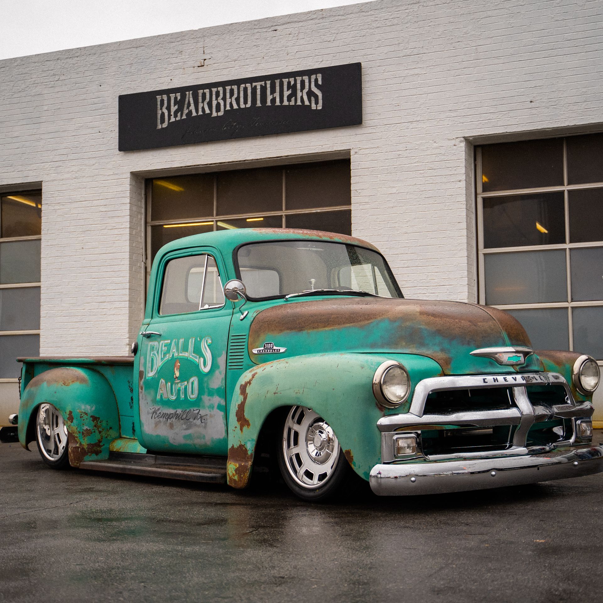 Rusty turquoise vintage pickup truck parked outside a BEARBROTHERS garage on a wet street