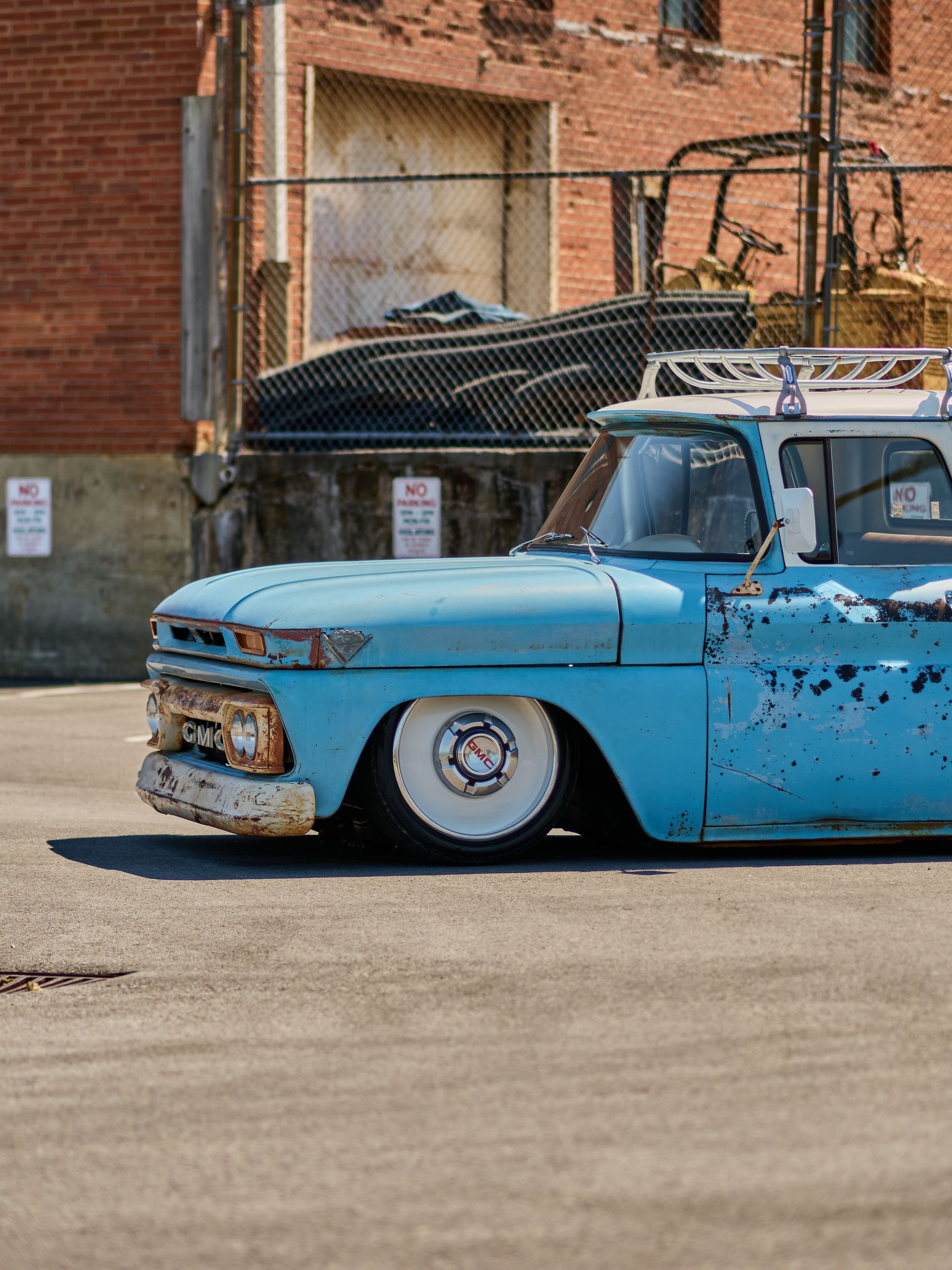 Blue lowered vintage pickup truck parked beside a brick wall and chain-link fence.