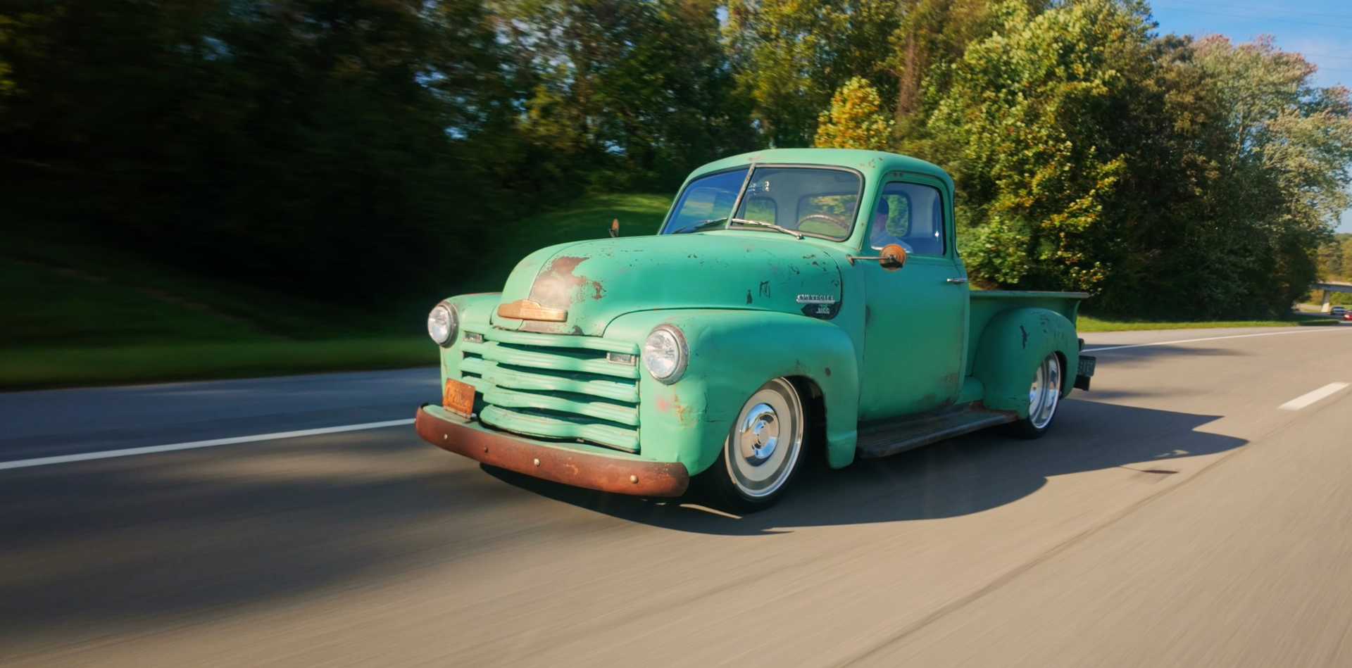 Mint-green vintage pickup truck driving on a sunlit road beside trees