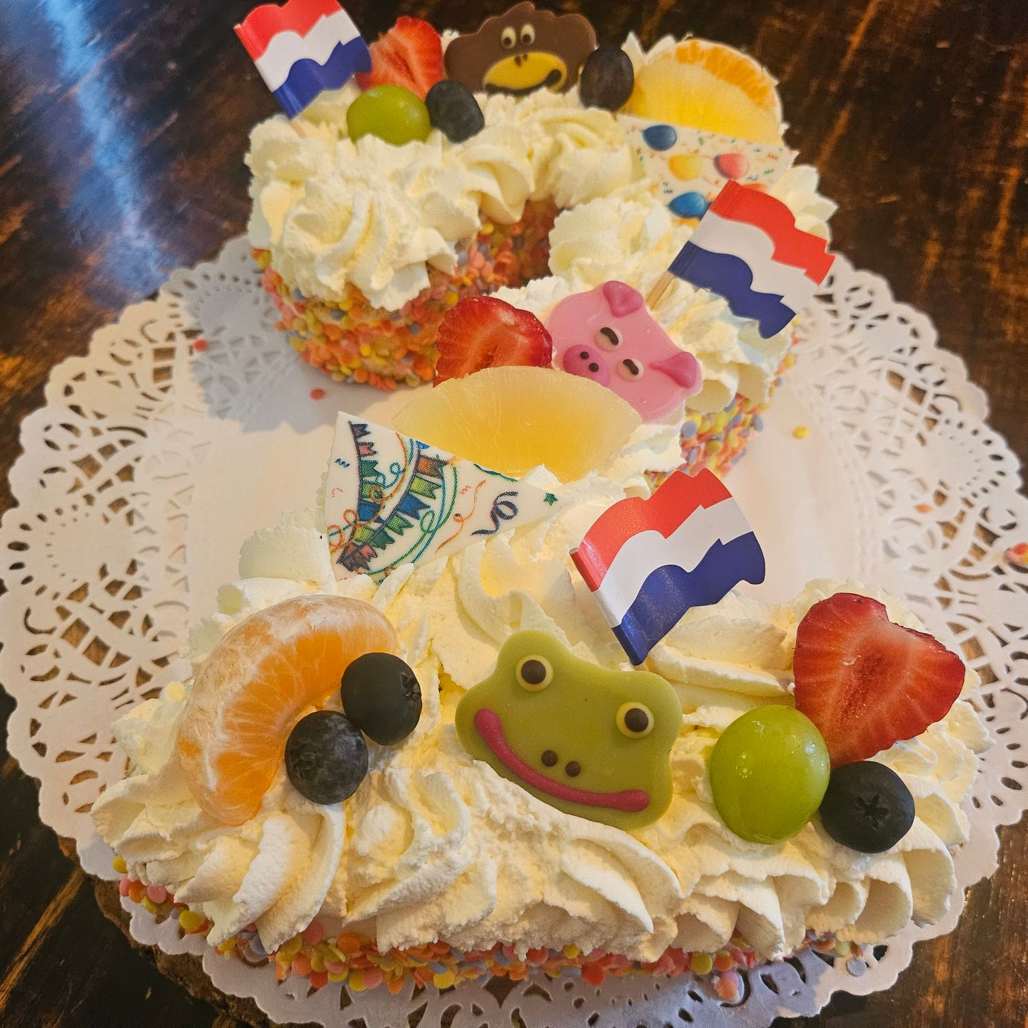 Versierde slagroomtaart met fruit op een kanten onderzetter, met kleine vlaggetjes en een kikkerdecoratie. In vorm van een 2.
