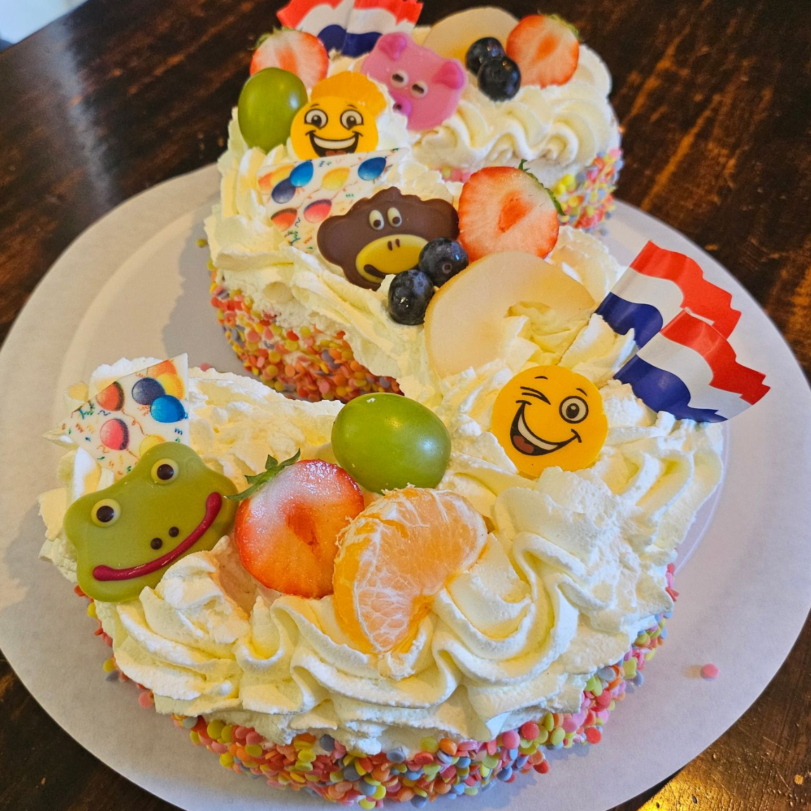 Versierde slagroomtaart met fruit op een kanten onderzetter, met kleine vlaggetjes en een kikkerdecoratie. In vorm van een 2.