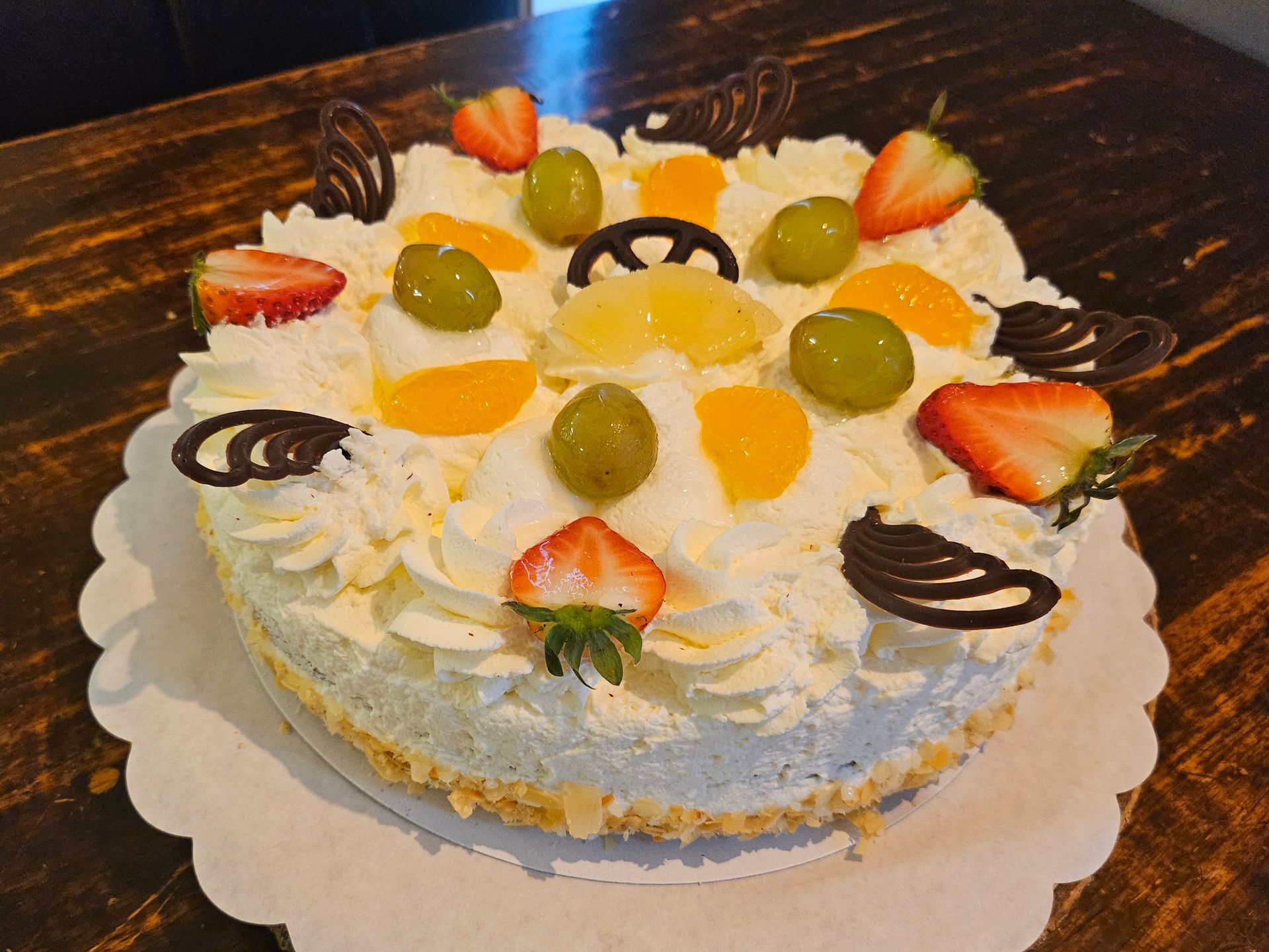 Ronde slagroomtaart met fruittopping op een gegolfd bord, versierd met aardbeien, kiwi, druiven en chocoladekrullen.