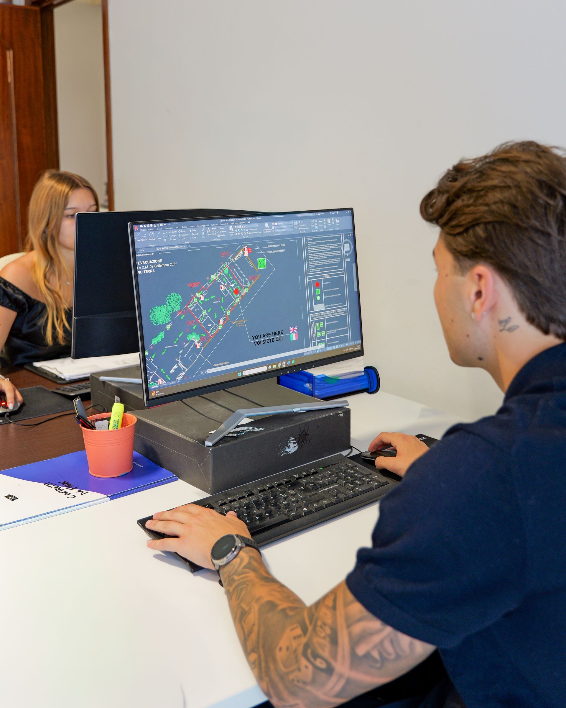 In un ufficio, due persone lavorano a una scrivania con un grande monitor che mostra una schermata simile a una mappa.