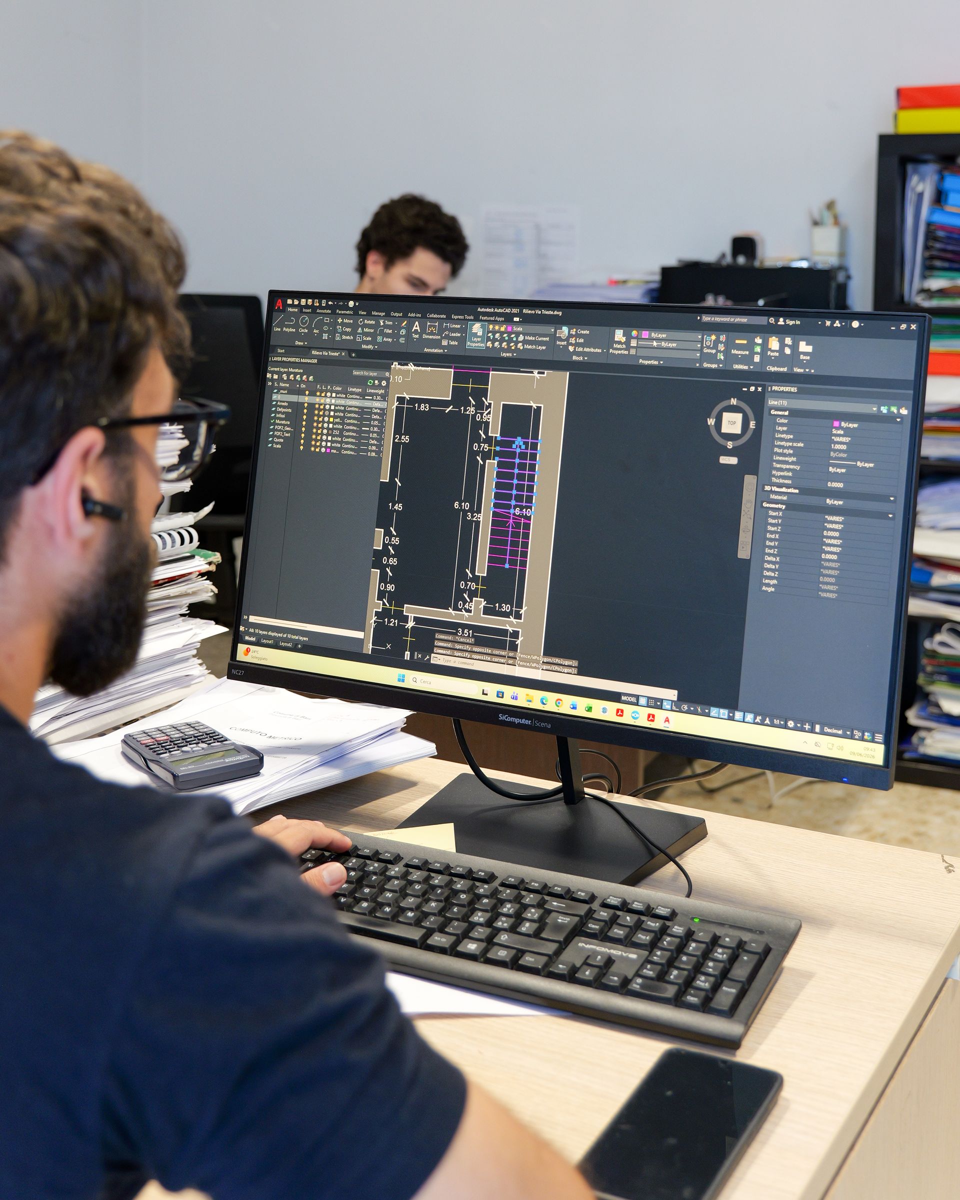 Una persona lavora al computer fisso in un ufficio condiviso, con del codice sullo schermo e un'altra persona sullo sfondo.