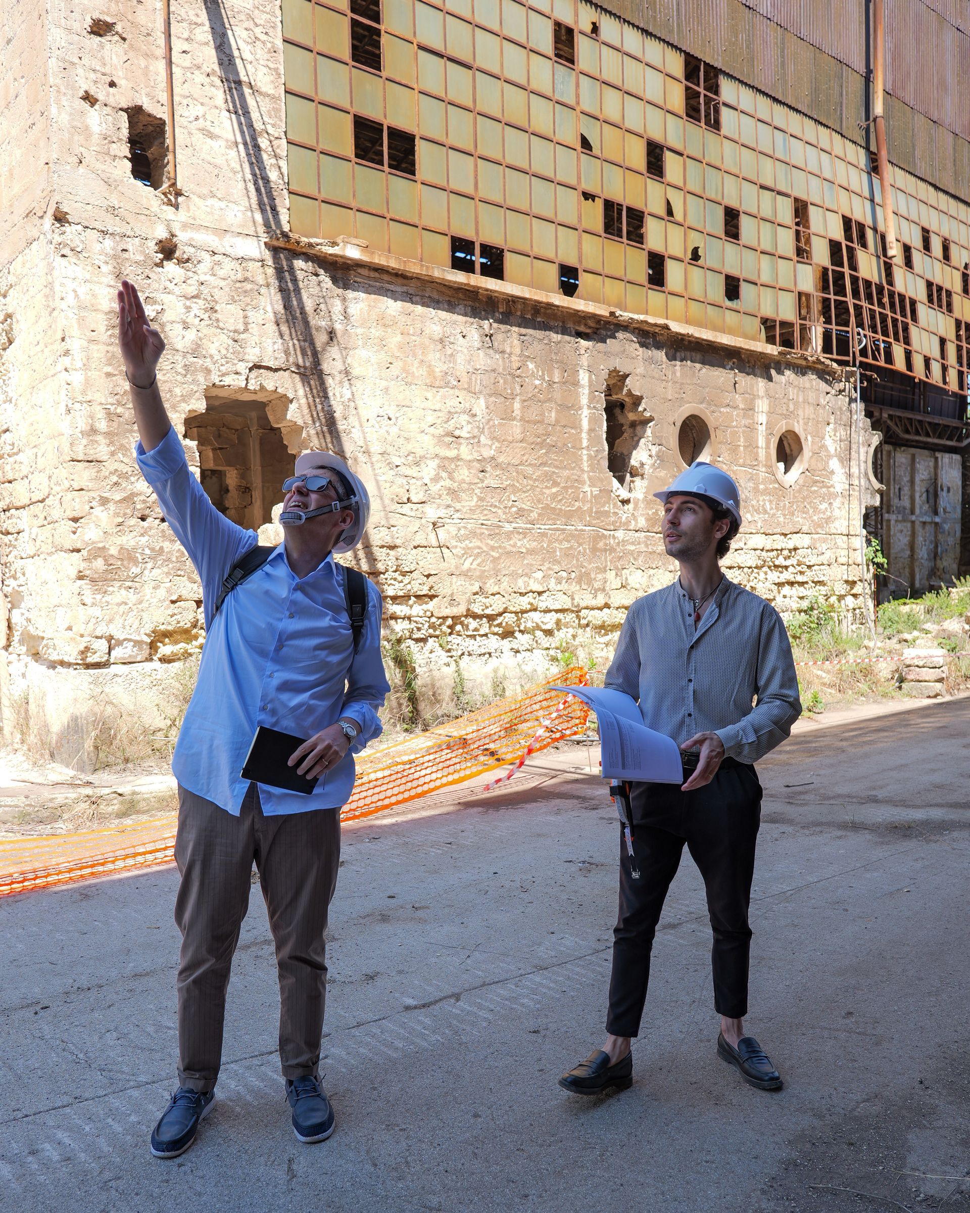 Due persone con l'elmetto sono in piedi accanto a un edificio industriale, una delle due indica verso l'alto con dei documenti in mano.