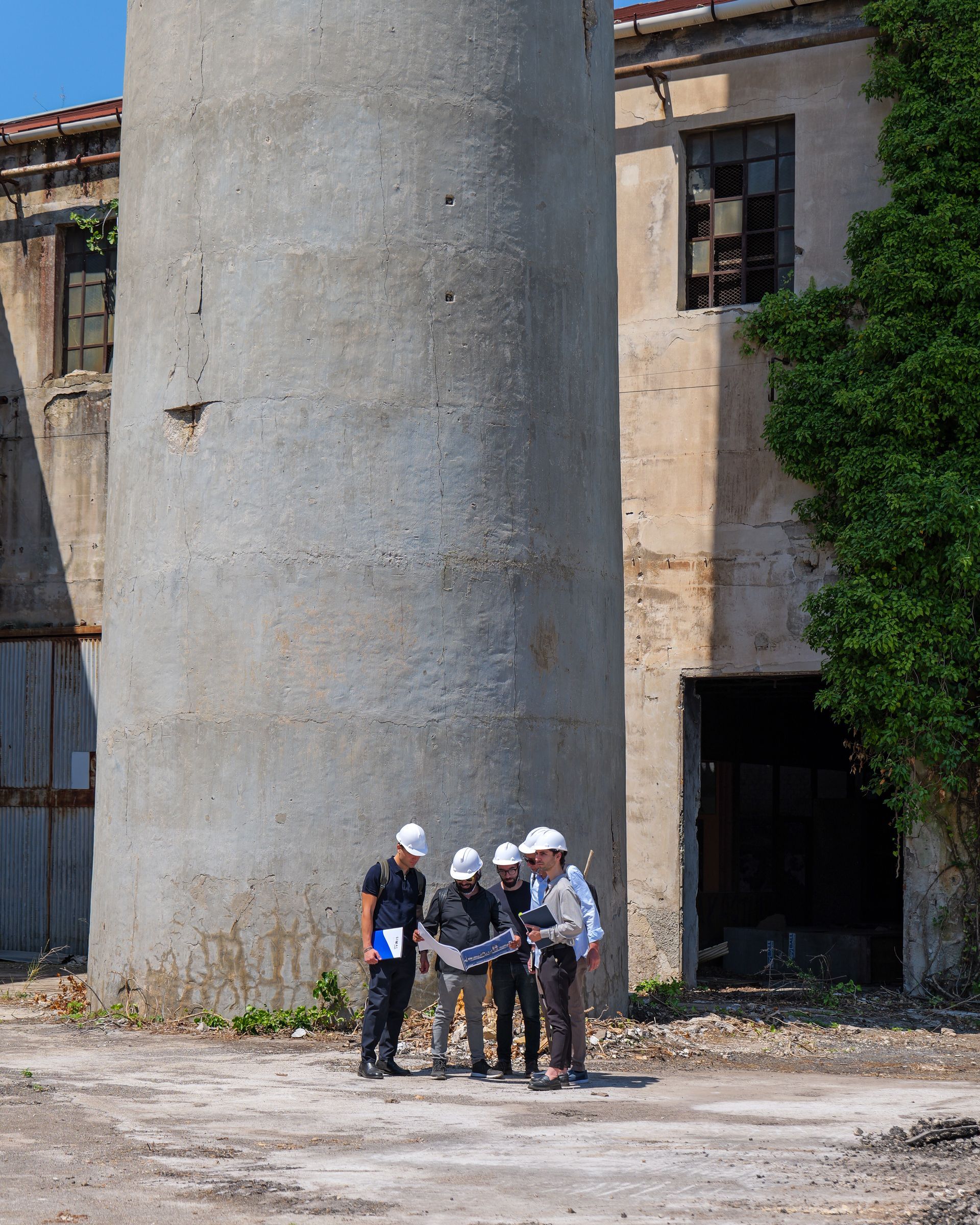 Quattro operai con elmetti di sicurezza ispezionano delle attrezzature accanto a un grande silo di cemento in un'area industriale.