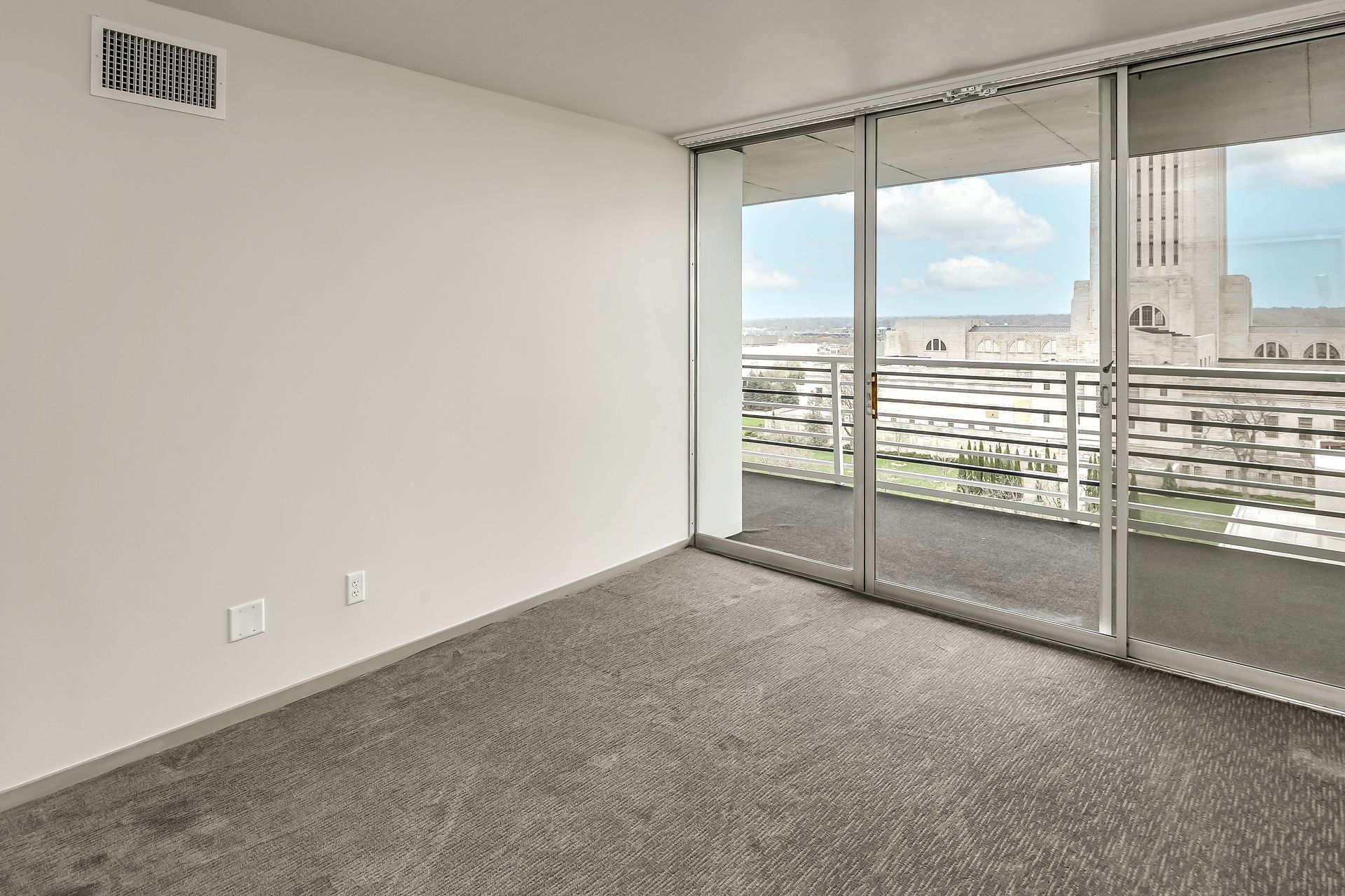 Empty carpeted room with sliding glass doors opening to a city view and balcony.