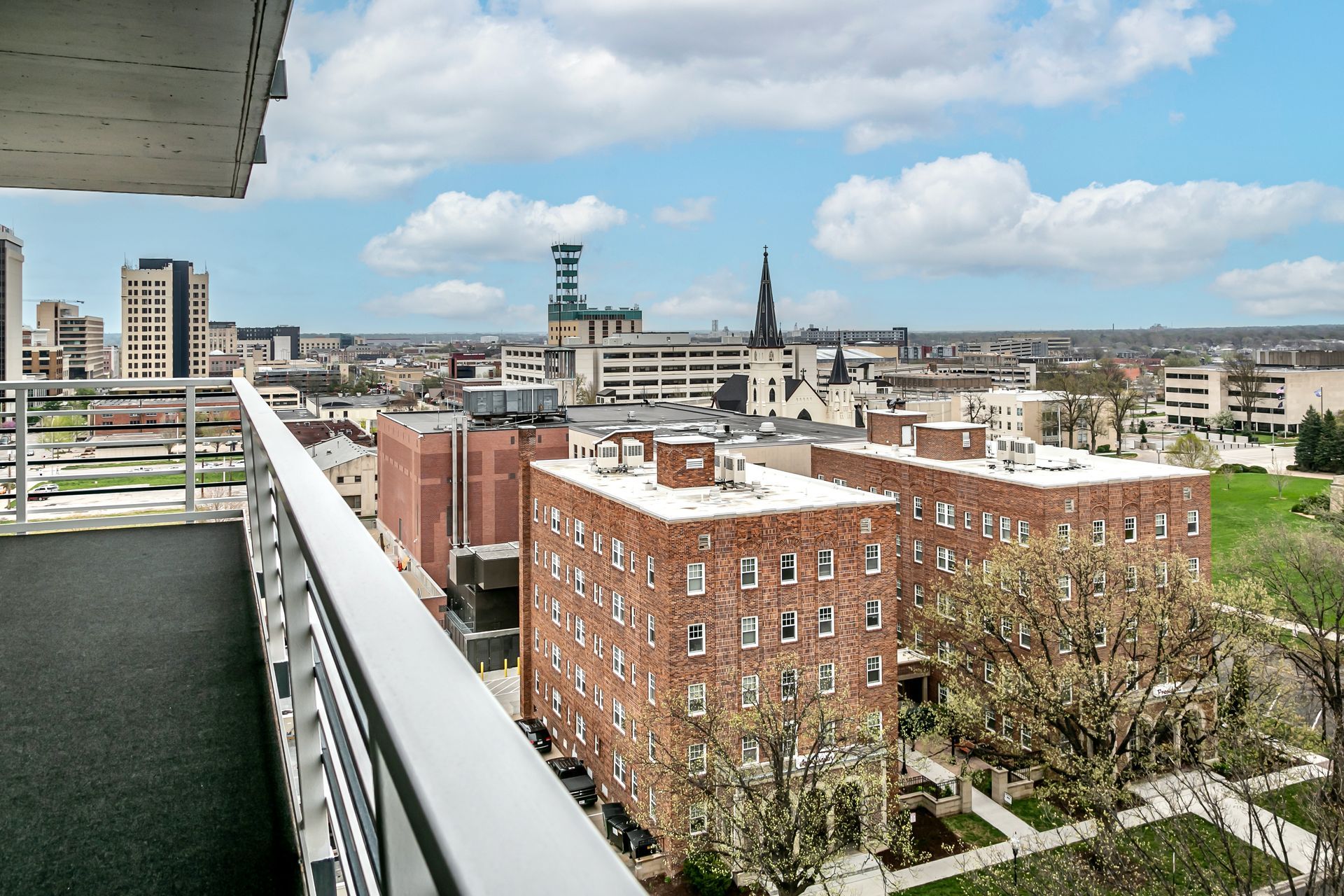 City skyline viewed from a balcony, with brick buildings, a church spire, and cloudy sky