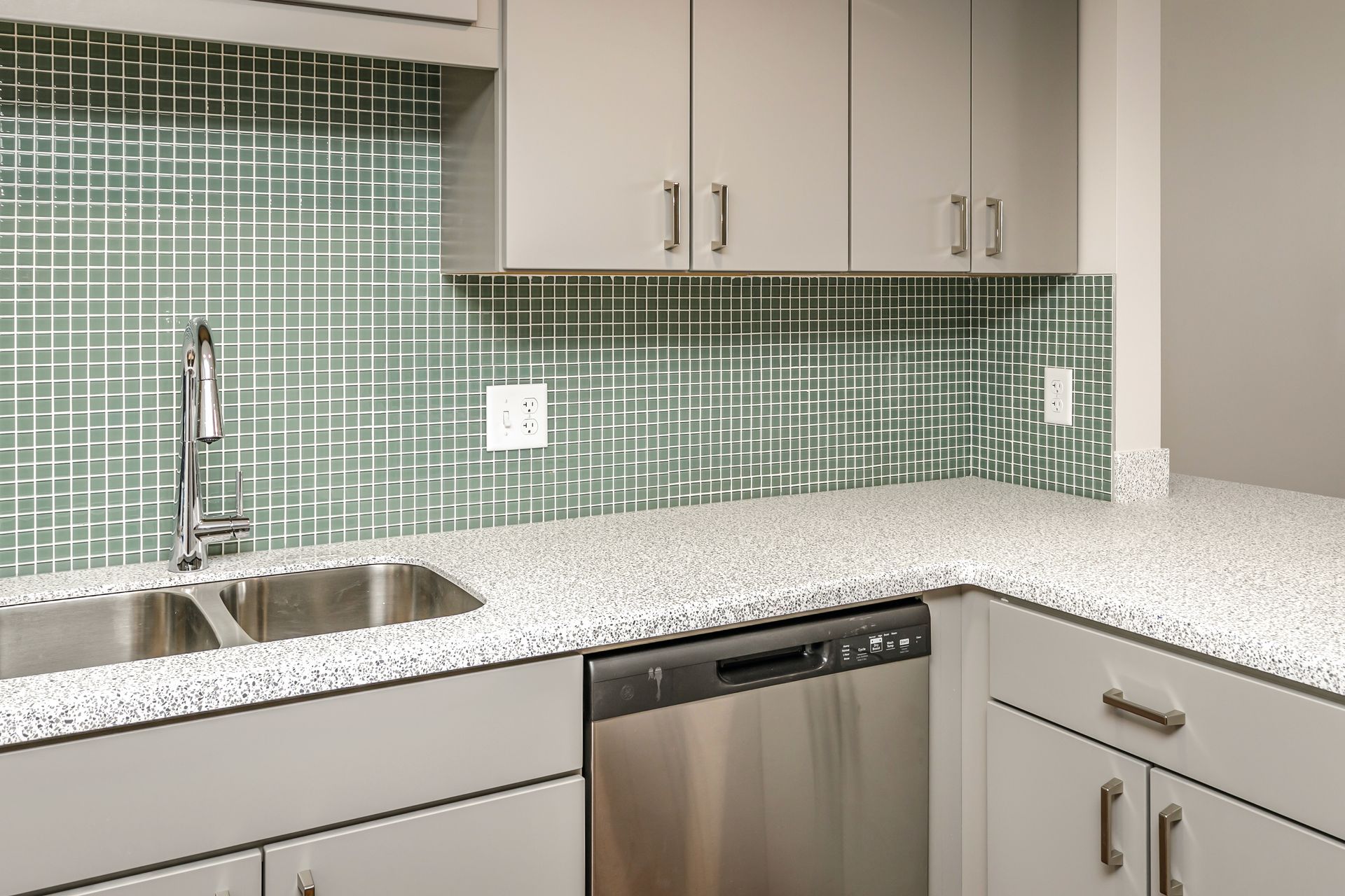 Modern kitchen corner with stainless sink, green tile backsplash, white countertops, and dishwasher