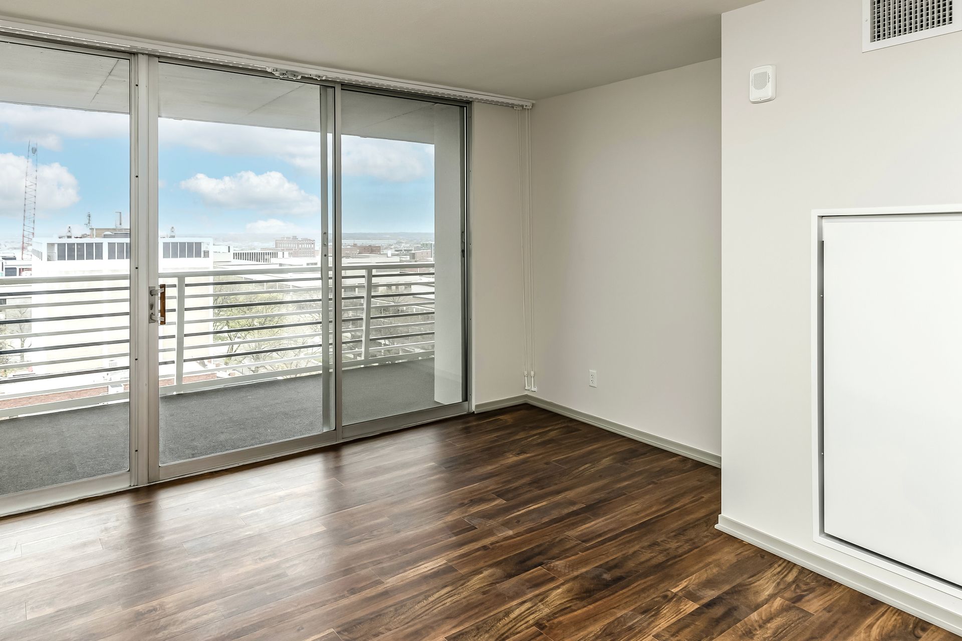 Empty room with dark wood floors, sliding glass doors, and a balcony view of buildings and sky.