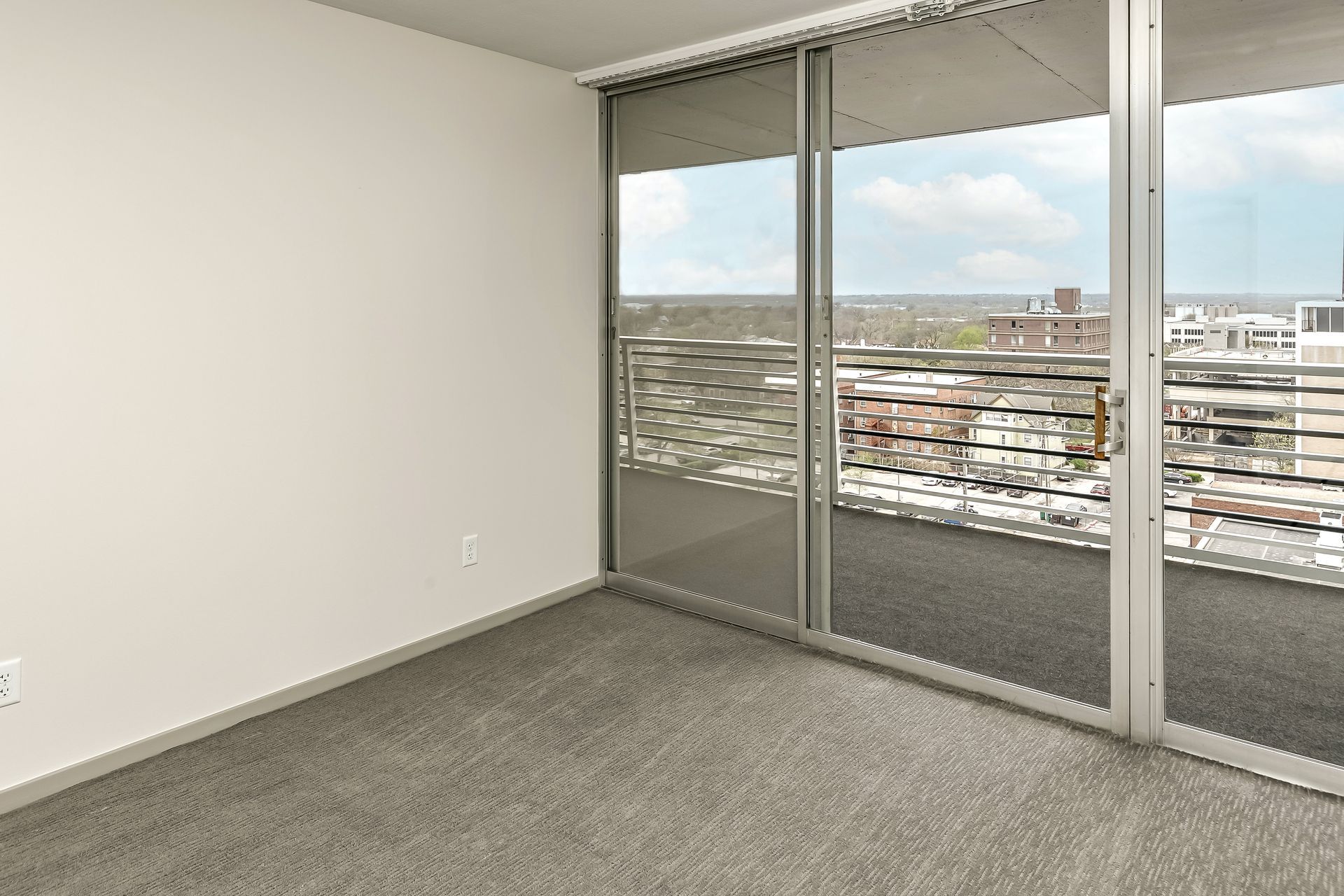 Empty carpeted room with large sliding glass doors opening to a city view.