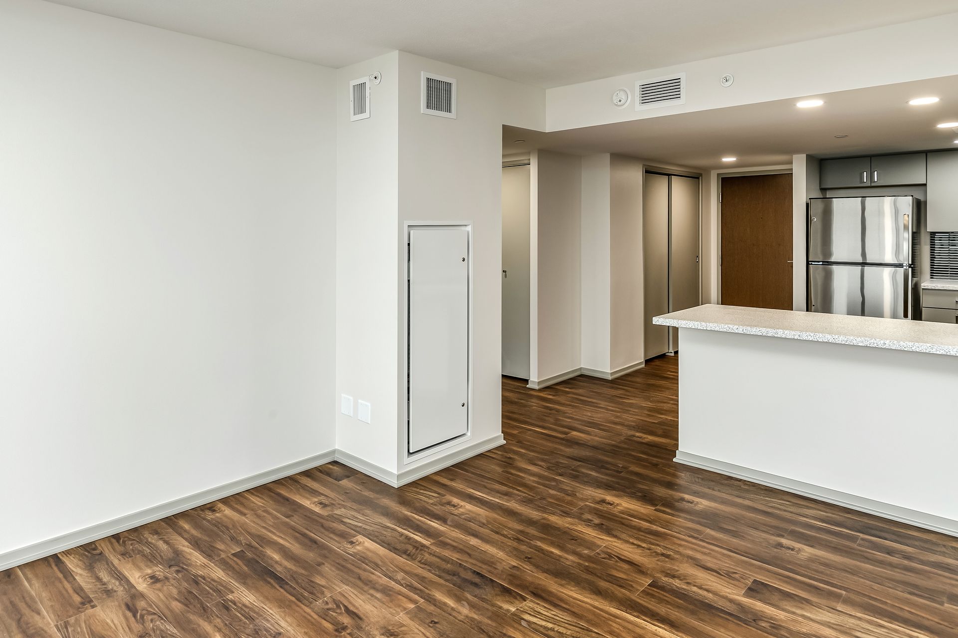 Empty modern apartment living area with wood floors, white walls, and a kitchen island view