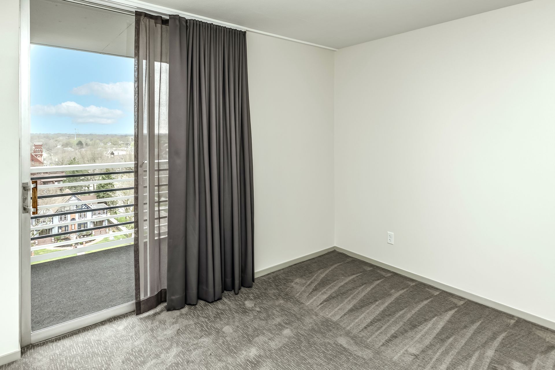 Empty gray-carpeted room with white walls and a sliding glass door with dark curtains.