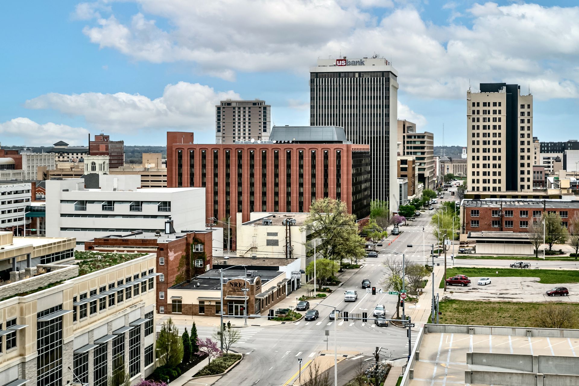Downtown city street with tall buildings, parked cars, and a partly cloudy sky