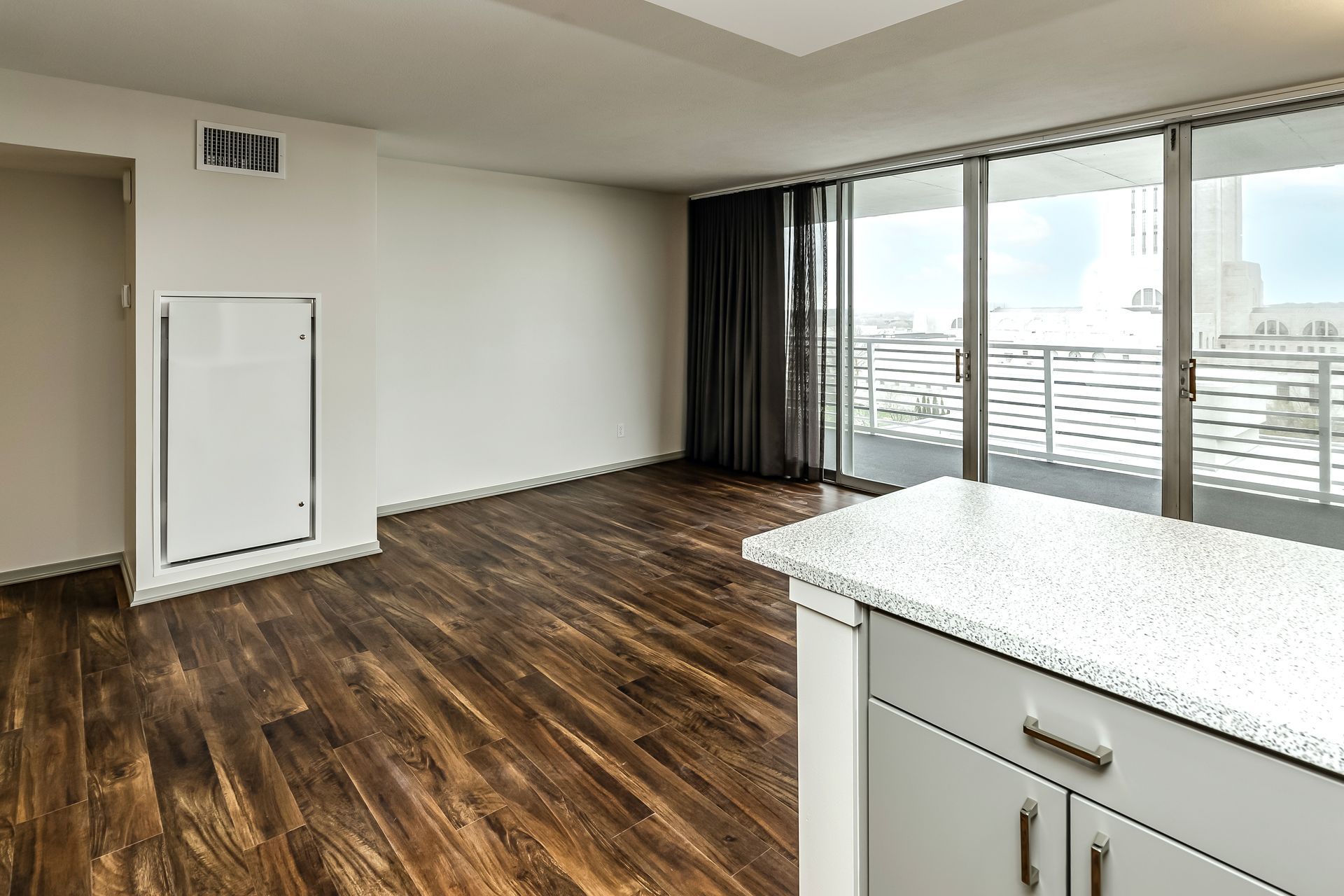 Empty modern living room with dark wood floors, white walls, and floor-to-ceiling windows overlooking a balcony