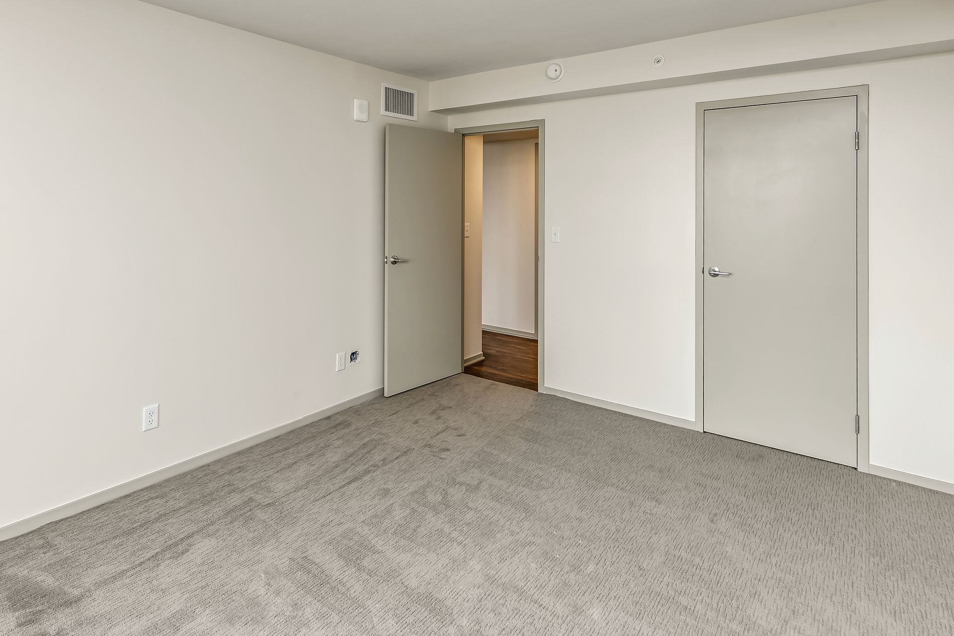 Empty carpeted room with white walls and two doors, one open to a hallway