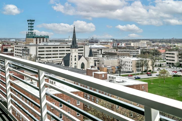 City skyline viewed from a balcony, with a white church tower and buildings under a partly cloudy sky.
