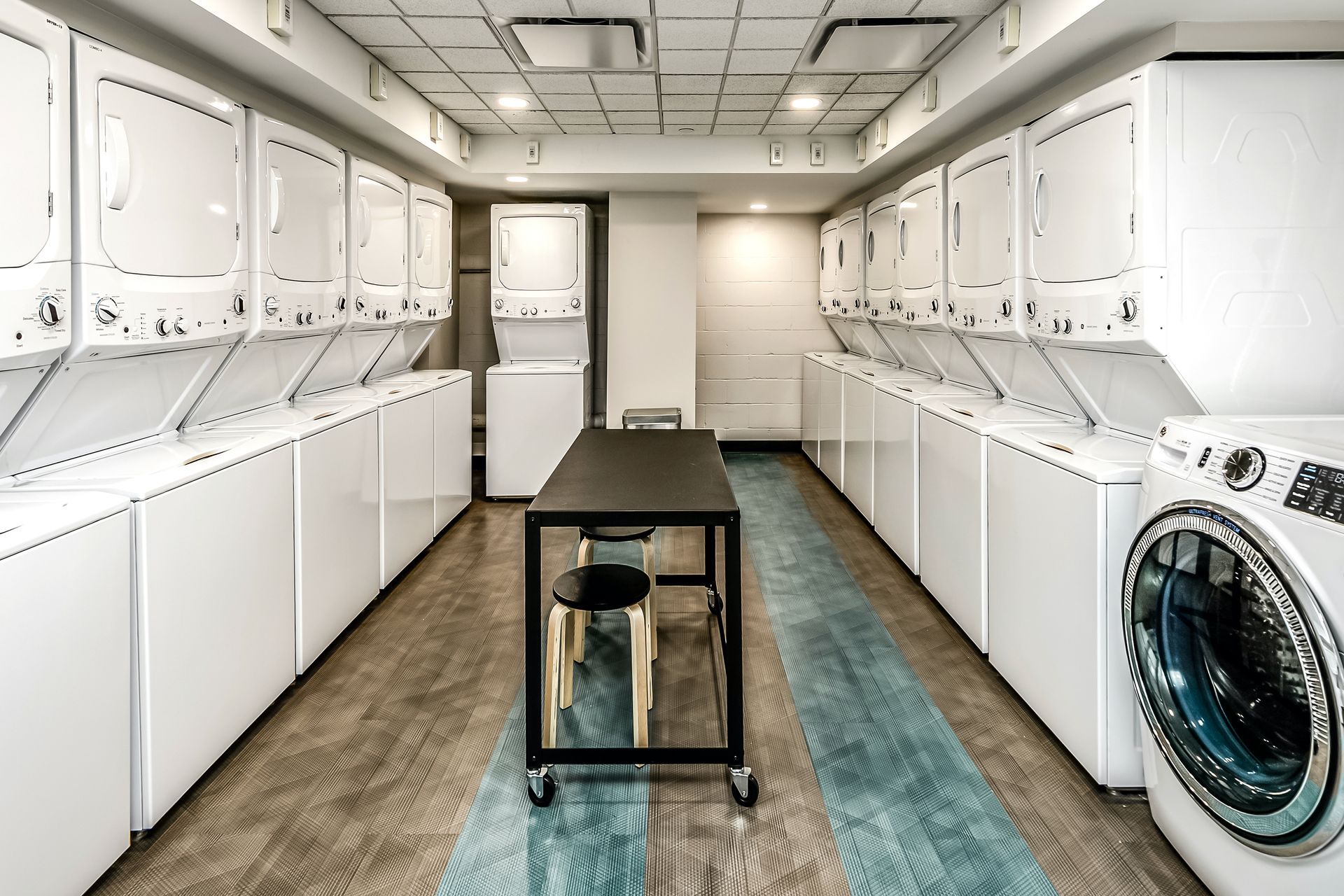 Modern laundromat with rows of white washers and dryers and a small table on a blue runner rug.