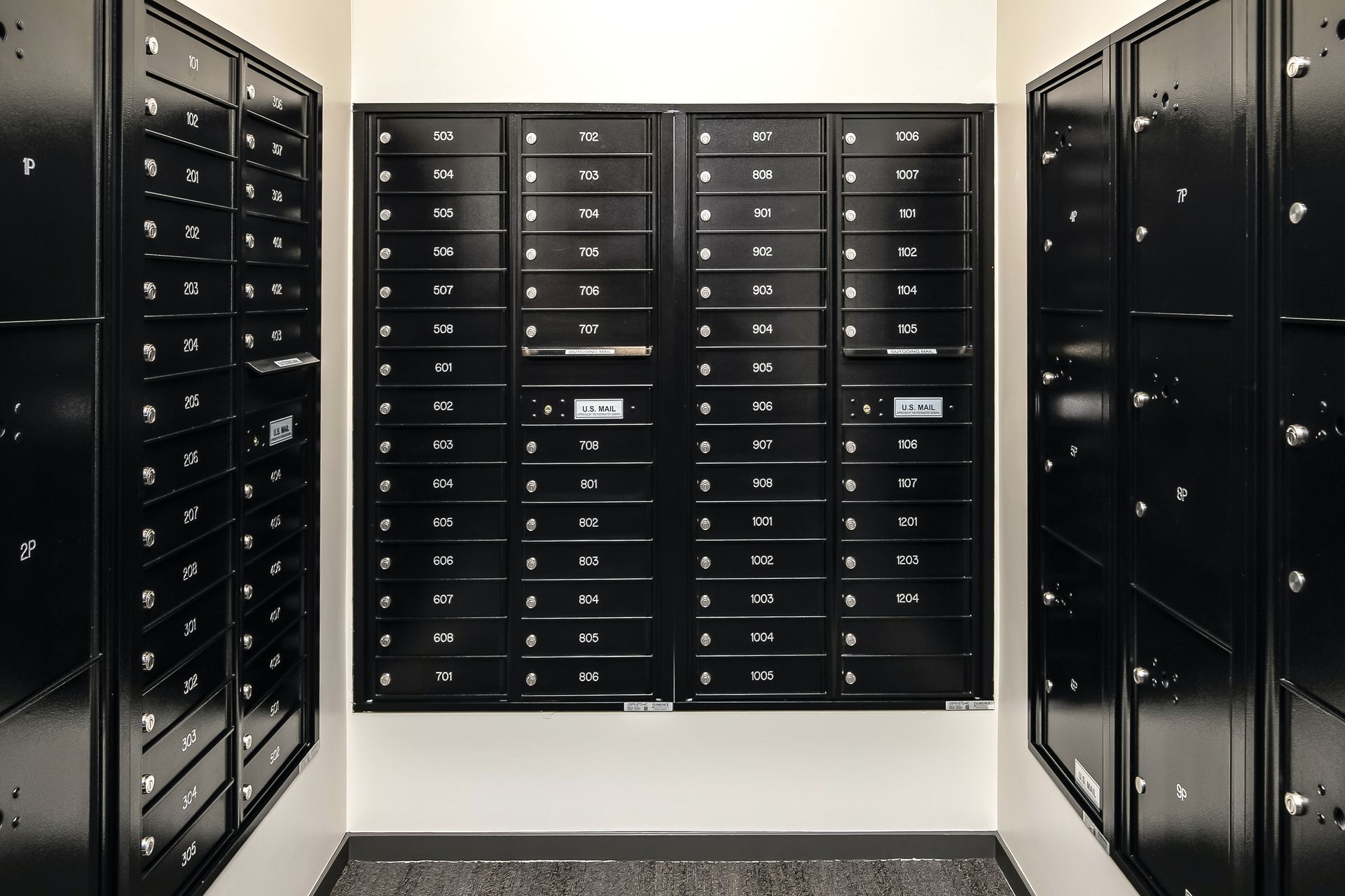Black mailboxes lined in rows inside a mailroom corridor with white walls and a tiled floor