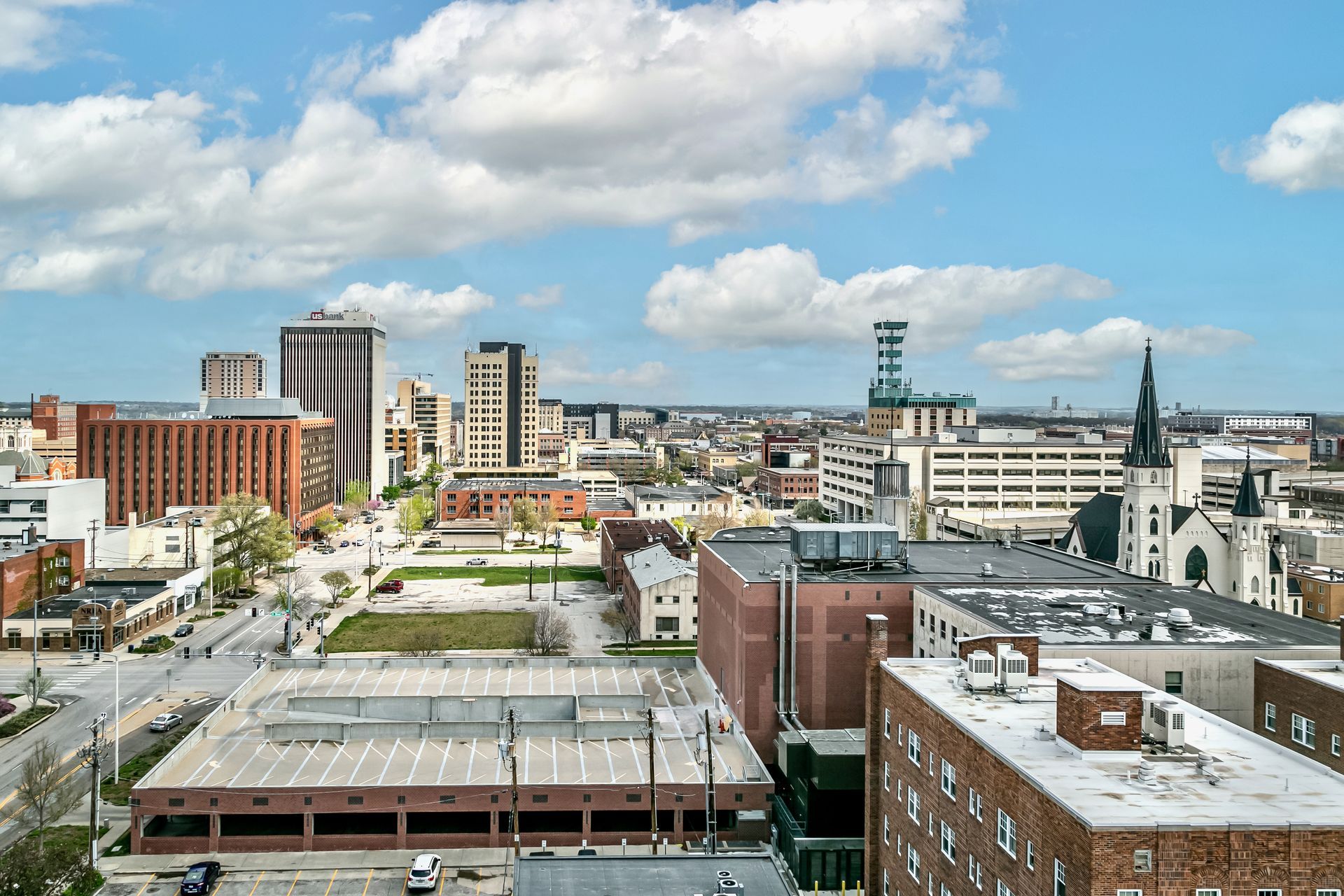 City skyline with mid-rise buildings, a church spire, and a cloudy blue sky.