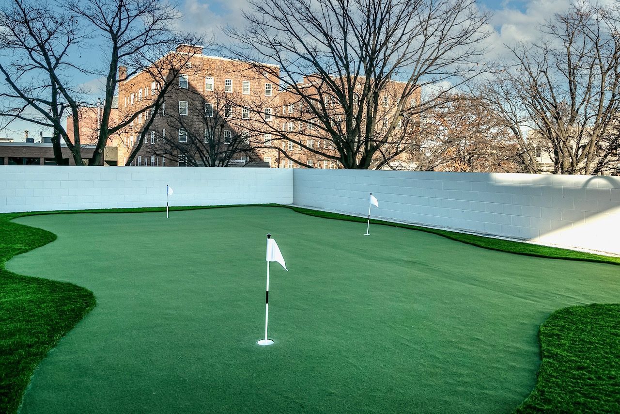 Small green putting green with white flag, surrounded by white wall and trees in the background