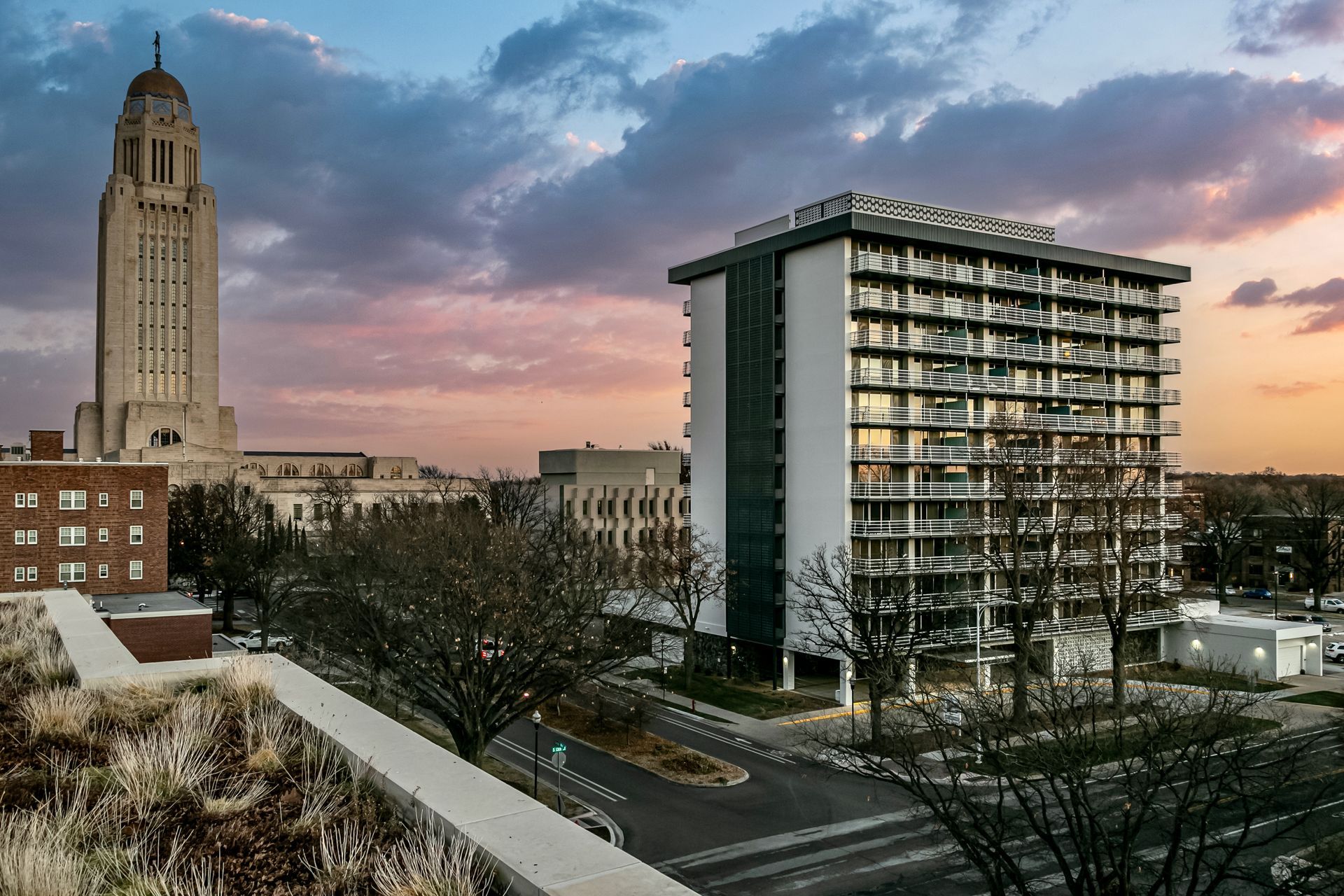 City skyline at dusk with a tall tower, modern apartment building, and cloudy pink-blue sky.
