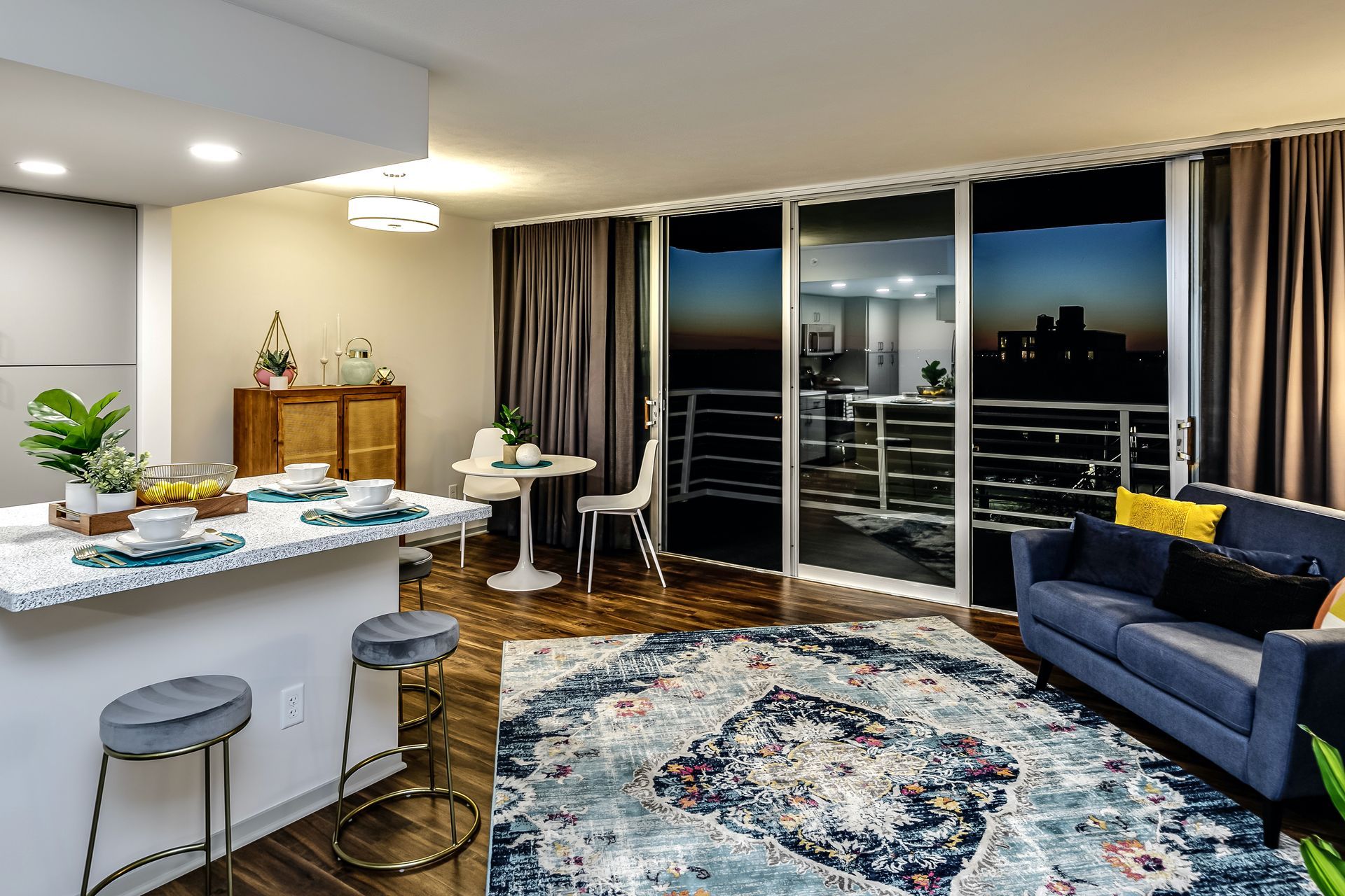 Modern living room with blue sofa, patterned rug, dining area, and sliding doors to a balcony at night